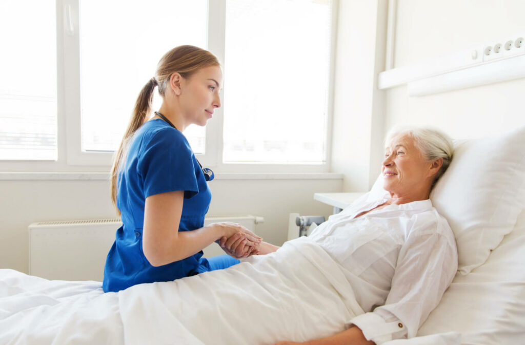 A female nurse in a blue scrub suit is sitting on the side of the bed while she is holding the hand of a female senior that is lying on the bed.
