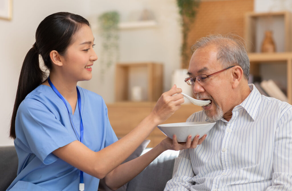 A young healthcare worker feeding a senior man in a striped shirt.