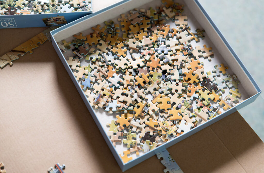 A box containing puzzle pieces sitting on top of a piece of cardboard where the outline of a puzzle is partly assembled